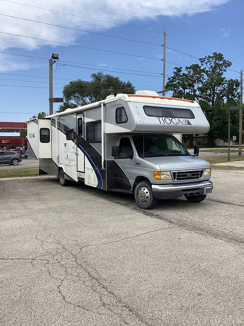 A white and gray fifth-wheel RV with black and blue graphics, parked on an asphalt lot under a blue sky. The RV has 'Tioga' written on the front overhang.